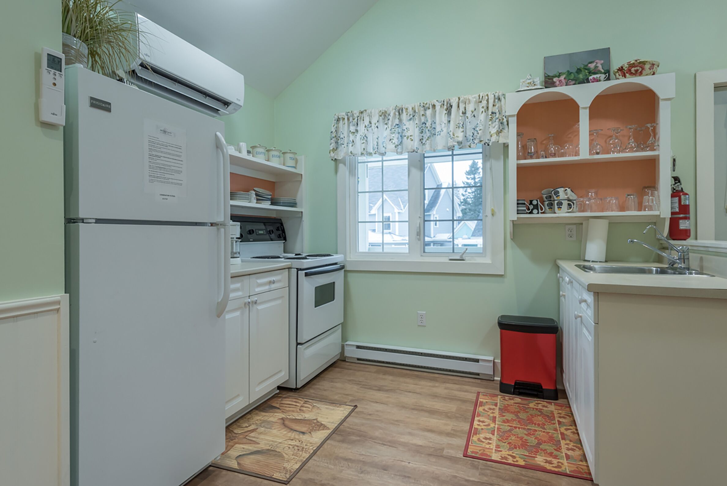 Uma cozinha com eletrodomésticos brancos, uma janela com cortinas florais e uma lixeira vermelha.