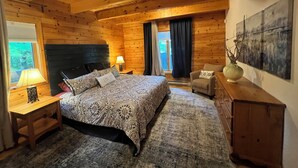 Ein gemütliches Schlafzimmer in einer Holzhütte mit Bett, Nachttisch, Lampe, Stuhl und Bildern an der Wand.