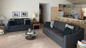 Een woonkamer met twee grijze banken, een glazen salontafel, en een keukenhoek met houten kastjes en een magnetron.