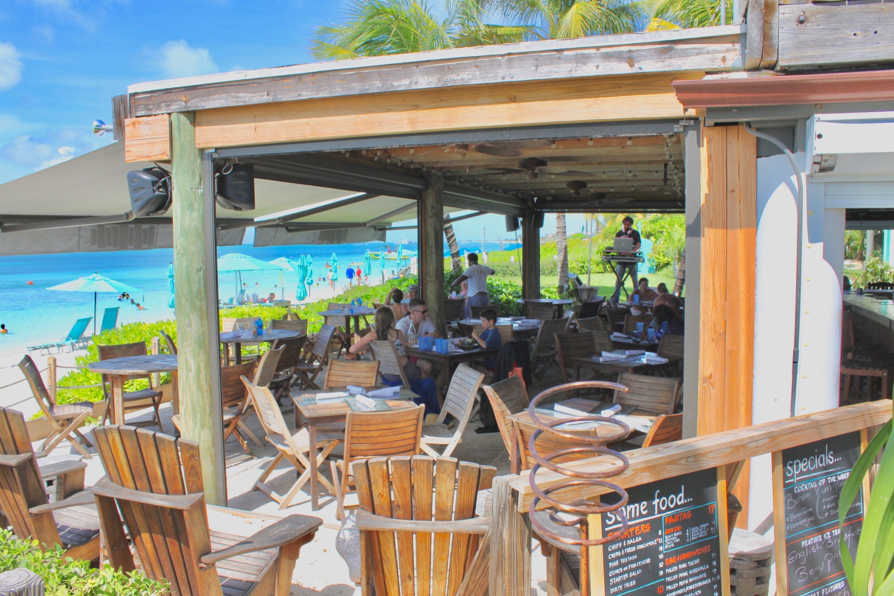 An outdoor seating area with wooden chairs and tables, overlooking a beach with umbrellas and people.