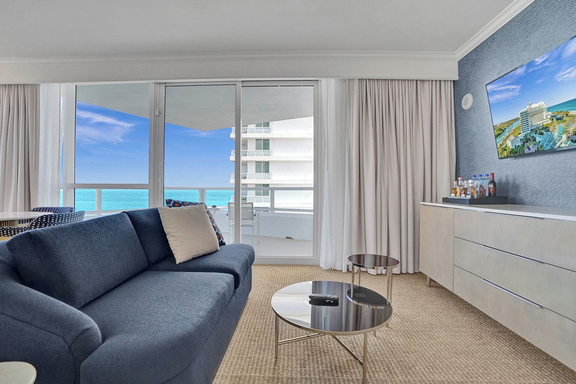 A hotel room with a balcony, a sofa, a small table, a minibar, and a large window with a view of the ocean and a building.