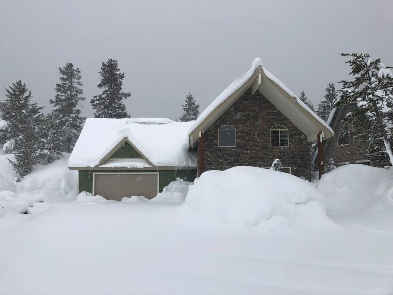 Una casa con tejado inclinado cubierto de nieve, rodeada de montones de nieve y árboles al fondo.