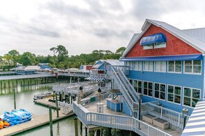 Un edificio frente al agua con techo rojo y detalles azules, amarrado en un puerto deportivo con barcos y una zona de muelles con asientos y sombrillas.