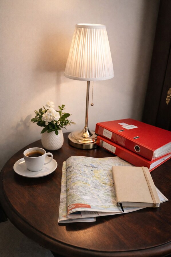 Une table ronde en bois, sur laquelle se trouvent une tasse à café blanche, un vase blanc avec des fleurs, une carte, un dossier rouge et une lampe.