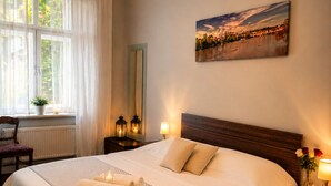 A neatly made bed with a patterned bedspread, a nightstand with a lamp, a window with sheer curtains, and a painting on the wall.