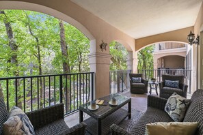 A covered outdoor seating area with wicker furniture, a glass table, and a view of a wooded area.