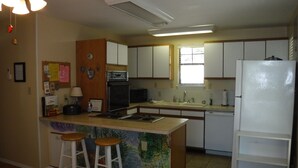 Una cocina con armarios blancos, horno empotrado, microondas, lavamanos y refrigerador. Hay dos taburetes en la isla y se ve un ventilador de techo.