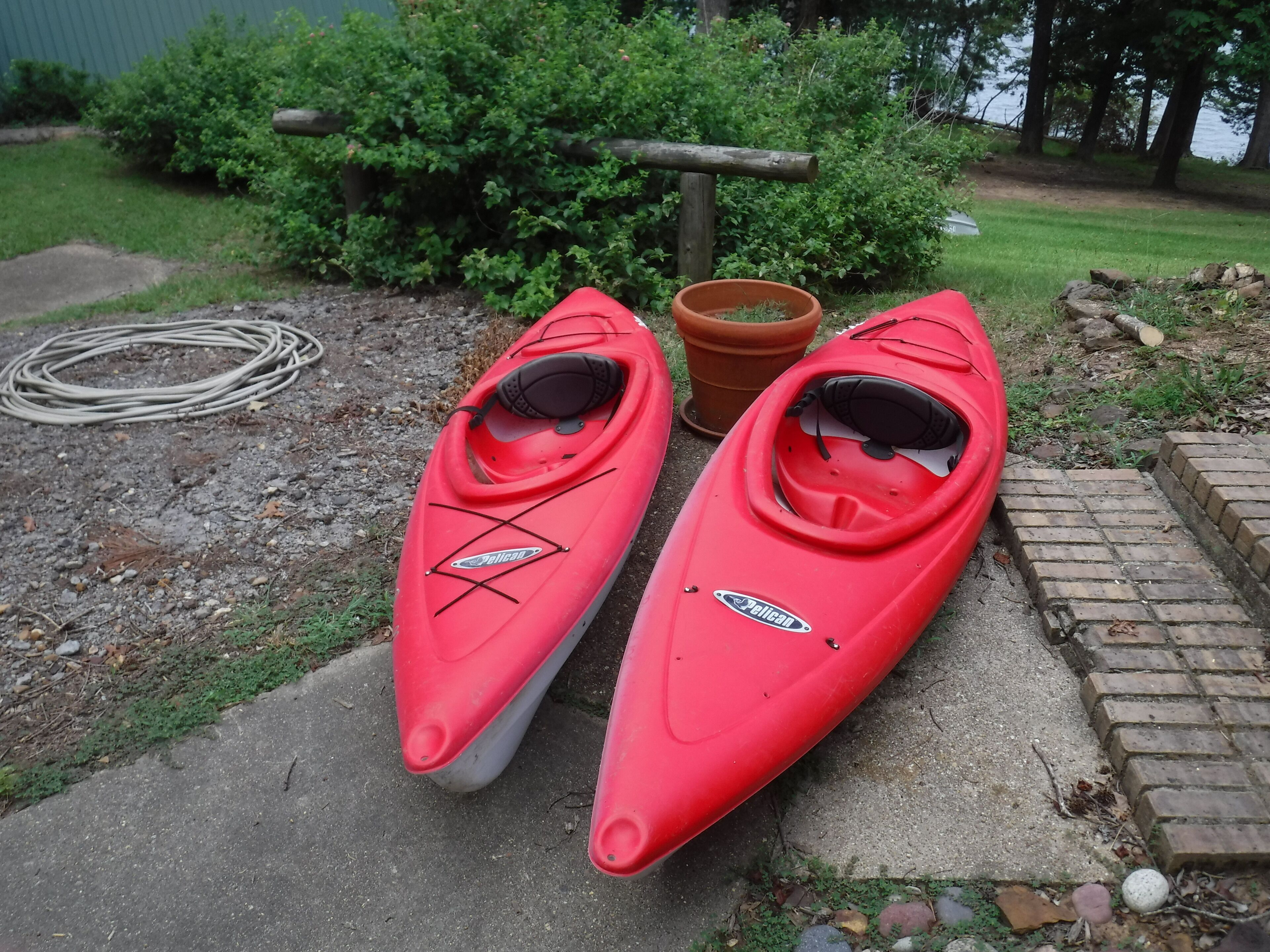 Dos kayaks rojos con el nombre de marca 'Pelican', colocados sobre una superficie de concreto al lado de un muro de ladrillo y un poste de madera.