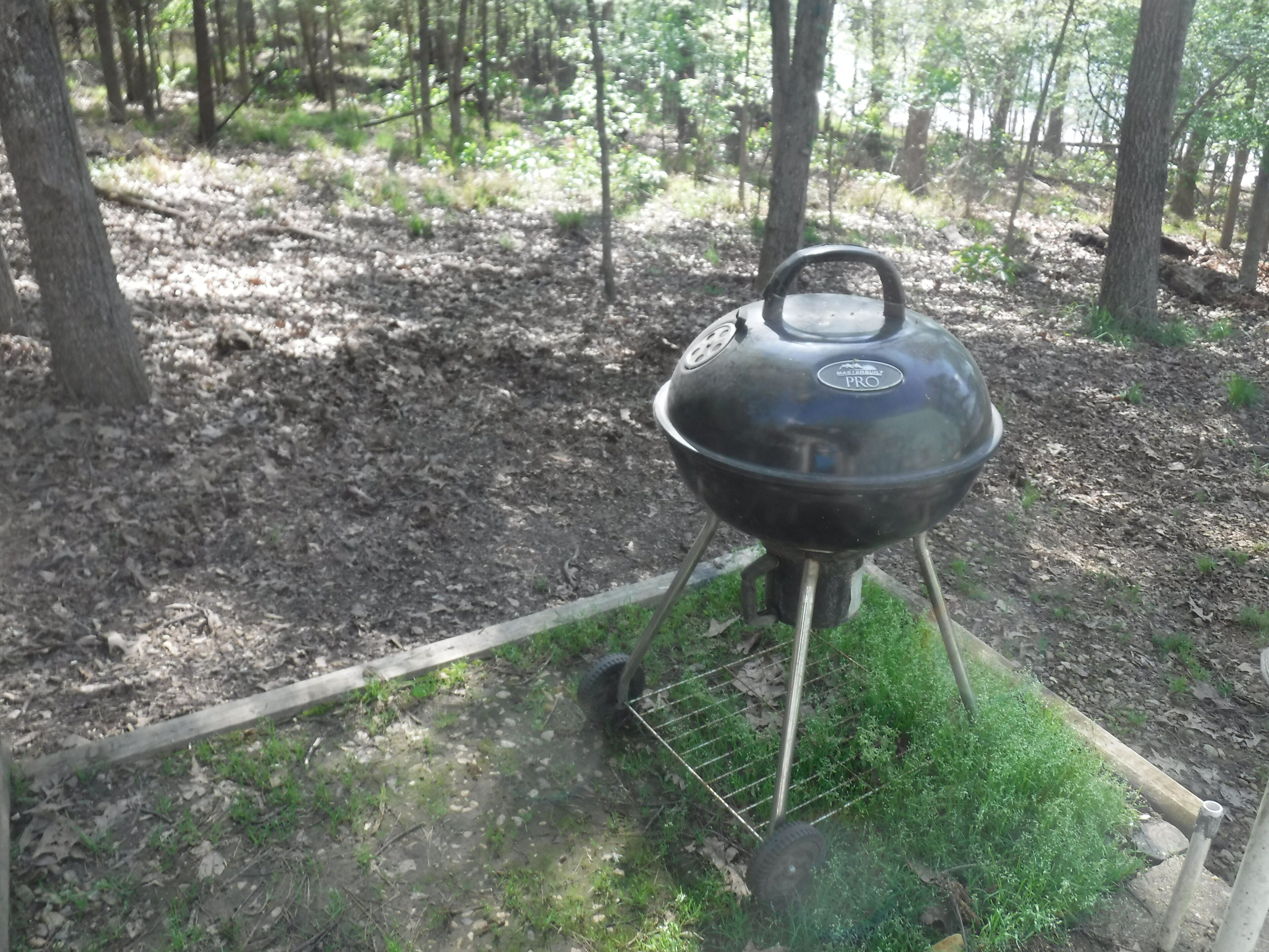 A portable barbecue grill with a lid and a handle, set up on a grassy area with a wire support.