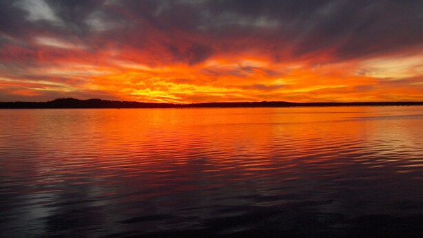 Ein Sonnenuntergang über ruhigem Wasser, im Hintergrund eine Silhouette von Land.