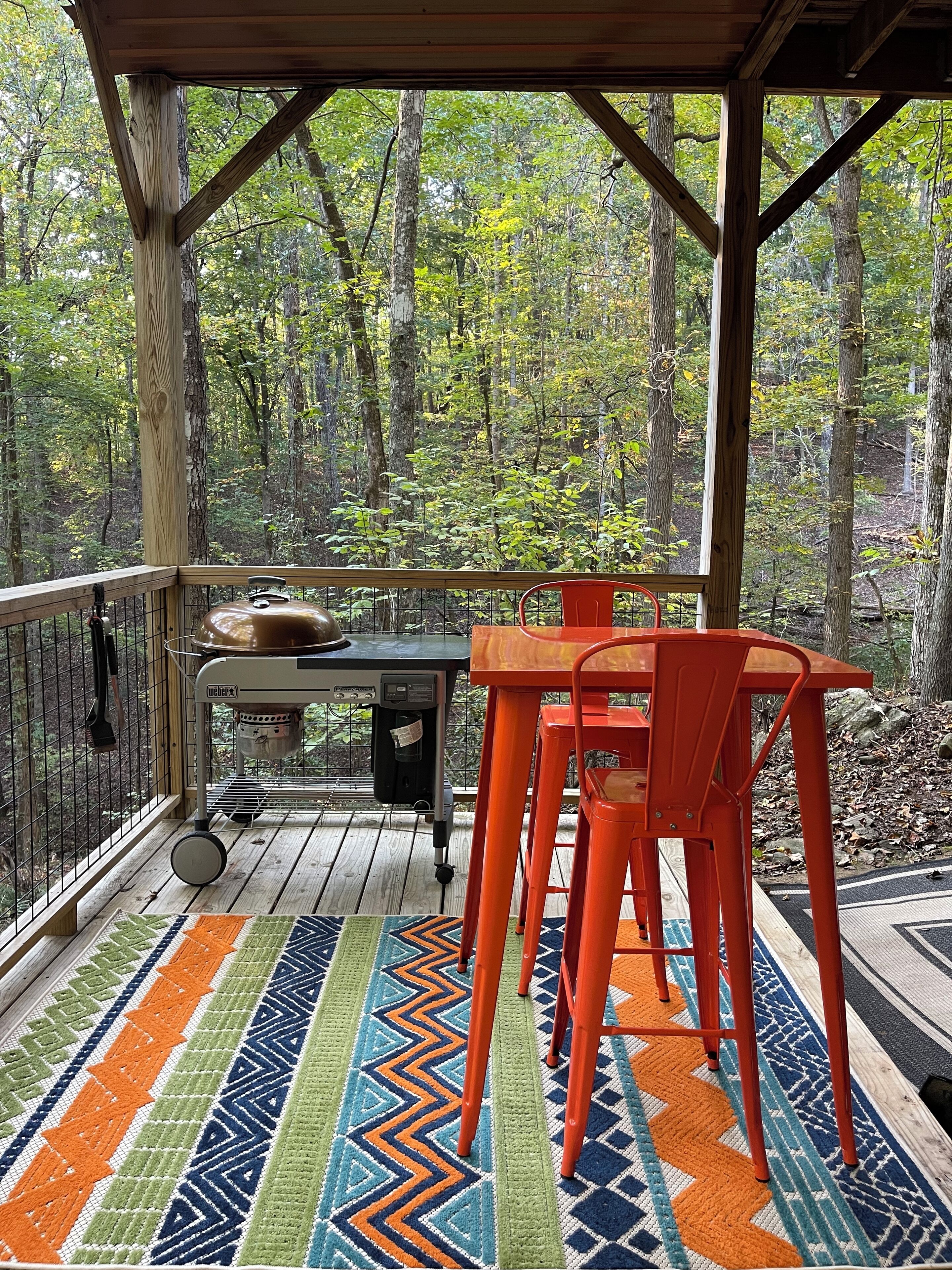Ein überdachter Außenbereich mit Tisch und Stühlen, umgeben von einer Holzterrasse und einem gemusterten Teppich.