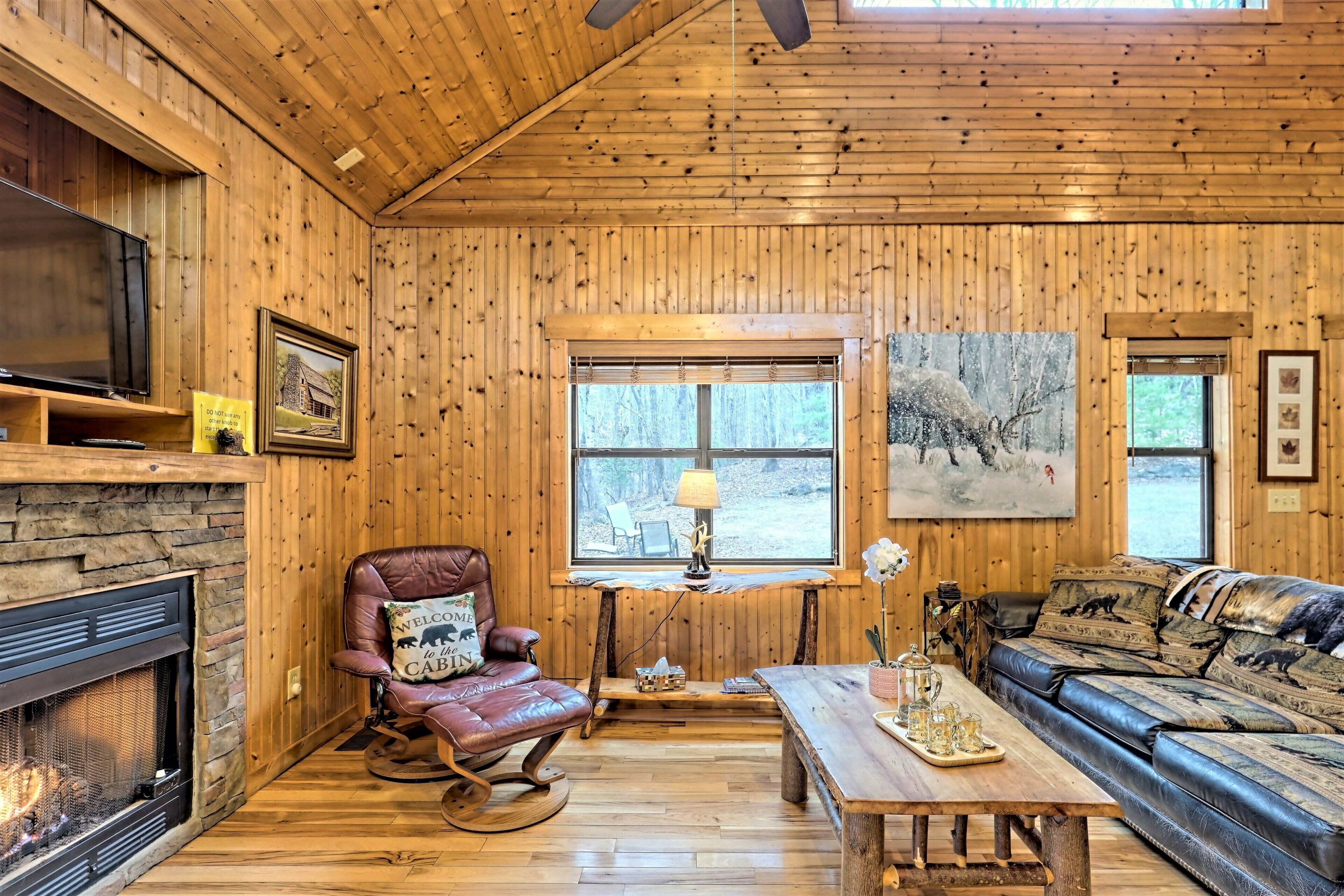A cozy cabin interior with a stone fireplace, leather armchair, wooden walls, and a large window overlooking a forest.