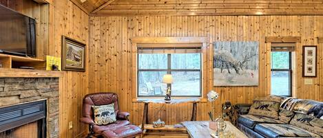 A cozy cabin interior with a stone fireplace, leather armchair, wooden walls, and a large window overlooking a forest.