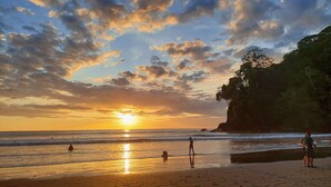 A beach with people and a dog near the water, a sunset, and a cliff with trees. - Gorgeous Views of Pacific and Rainforest (Manuel Antomio)