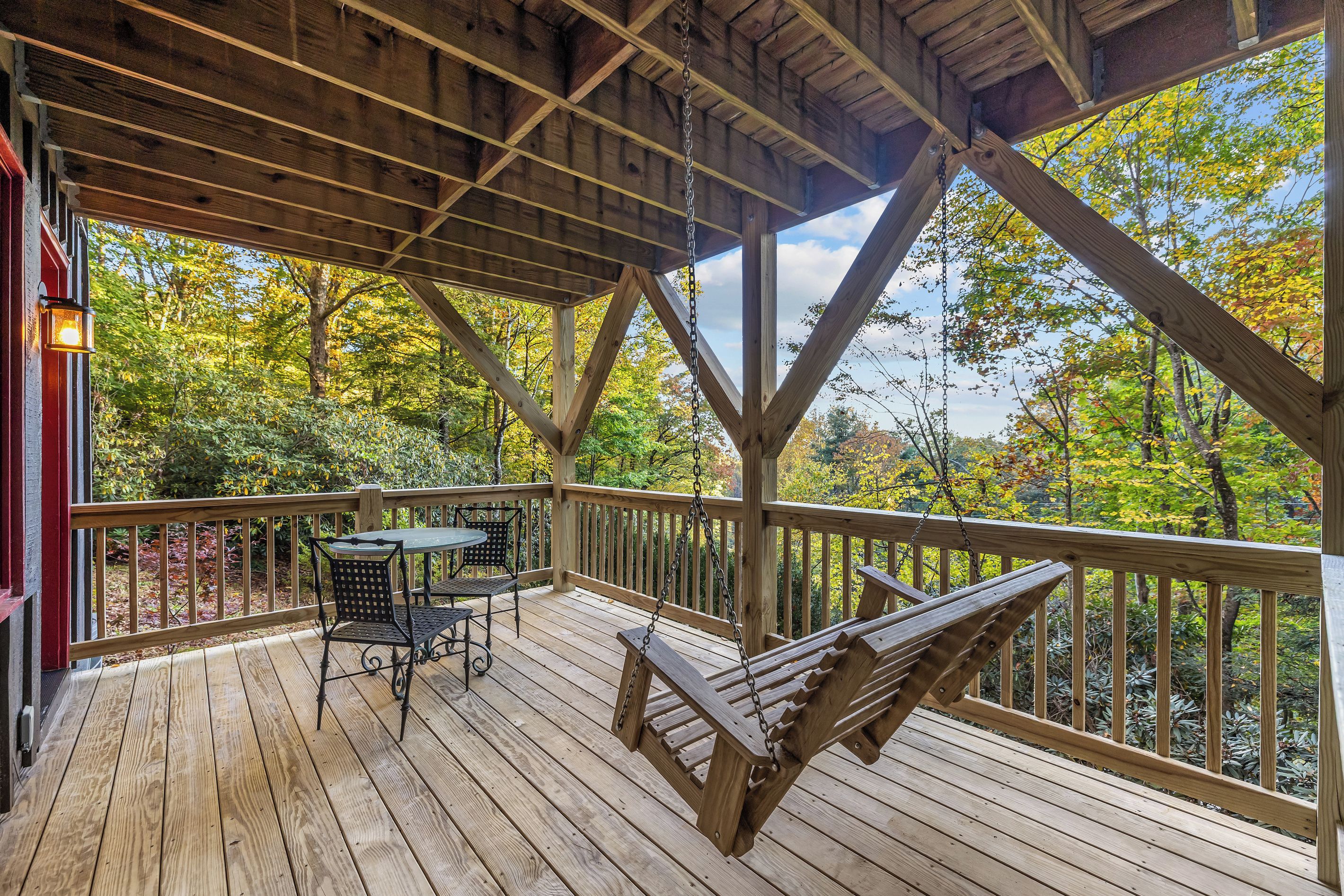 Une terrasse en bois avec une table et des chaises, une balançoire suspendue et une vue sur le feuillage automnal.
