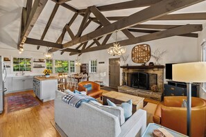 A spacious living area with a fireplace, wooden beams, and a dining area with a view of greenery outside.