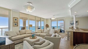 A spacious living room with a large sectional sofa, a coffee table, and a dining area with a bar. Large windows offer a view of the ocean. - Beach Club 1406 - 𝑺𝒆𝒂𝒔𝒊𝒅𝒆 𝑺𝒆𝒓𝒆𝒏𝒊𝒕𝒚 (Pensacola Beach)