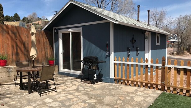A small, single-story building with a blue exterior and a white picket fence. It features a patio with stone tiles and a wooden table and chairs. - Newly Remodeled  Pet Friendly Cottage (Colorado Springs)