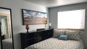 A bedroom with a bed, a bedside table, a lamp, a framed picture on the wall, and a window with blinds. - Newly Remodeled  Pet Friendly Cottage (Colorado Springs)