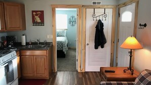 A compact kitchen with wooden cabinets, a stove, and a coffee maker. There is a small dining area with a table and a lamp. A bedroom is visible through an open door, featuring a bed and a hanging coat rack. - Newly Remodeled  Pet Friendly Cottage (Colorado Springs)