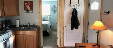 Una cocina compacta con armarios de madera, estufa y cafetera. Se observa un pequeño comedor con una mesa y una lámpara. Se puede ver un dormitorio a través de una puerta abierta, que incluye una cama y un perchero.