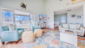 A spacious living room with a large window, a ceiling fan, and a colorful rug. - Oceanfront Dog Friendly Home in Waves w/ Private Pool, Hot Tub, Tiki Bar, Grill (Waves)
