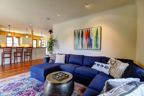 A living room with a blue sofa, a coffee table, and a colorful rug. There is a kitchen in the background with wooden cabinets and a dining area with chairs.