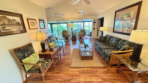 A spacious living room with wooden flooring, two sofas, a coffee table, and a chair. There are ceiling fans, framed pictures on the walls, and a view of greenery through large windows. - Paradise Found:  Spacious, Comfortable, Private Hawaiian Cottage (Lahaina)
