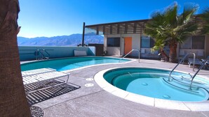 A pool area with a view of mountains, a palm tree, and a building with a covered deck. - The Getaway 1BR #2: Pool, Mountain View, Kids/Pets welcome (Desert Hot Springs)