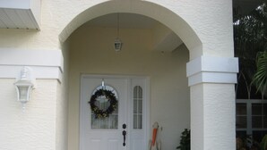 A white door with a wreath, a small white table with a sculpture, and a black doormat.
