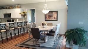 A dining area with a wooden floor, a dining table with chairs, a chandelier, a framed picture on the wall, and a potted plant.