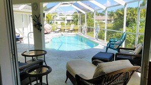 A covered outdoor area with a pool, wicker furniture, and a view of the surrounding greenery.