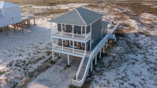 Una casa de playa de dos pisos con balcón y terraza, rodeada de arena y vegetación escasa.