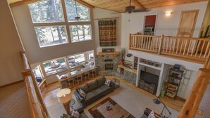 A spacious living area with a stone fireplace, a wooden balcony, and large windows offering a view of the outdoors. - Kantor's Hideout: Ample TD Home with Private Hot Tub, Pool Table, Walk to Trails (Truckee)