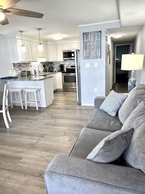 A modern kitchen with white cabinets, a granite countertop, and stainless steel appliances. There is a gray sofa with patterned cushions in the living area.