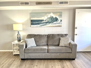 A grey sofa with patterned cushions, a lamp, and a painting of a wave on the wall.