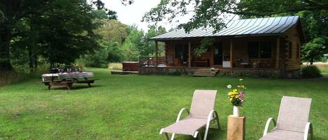 A rustic cabin with a porch, a picnic table, and two lounge chairs in a grassy area.