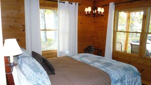 A wooden cabin bedroom with a bed, curtains, a chandelier, and a view of the outdoors. - Simple.  Rustic.  Luxury. (Monterey)