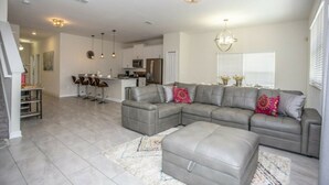 A spacious living room with a grey sectional sofa, a matching ottoman, and a kitchen in the background. - Astonishing Home Private Pool 4830 (Kissimmee)