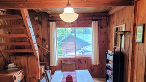 Ein Holzblockhaus-Interieur mit einem Esstisch, Stühlen, einer Treppe, einem Bücherregal und einem Fenster mit Blick auf ein Auto und Grünflächen.