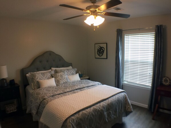 A bedroom with a bed, a ceiling fan, a window with blinds, and a nightstand with a lamp.