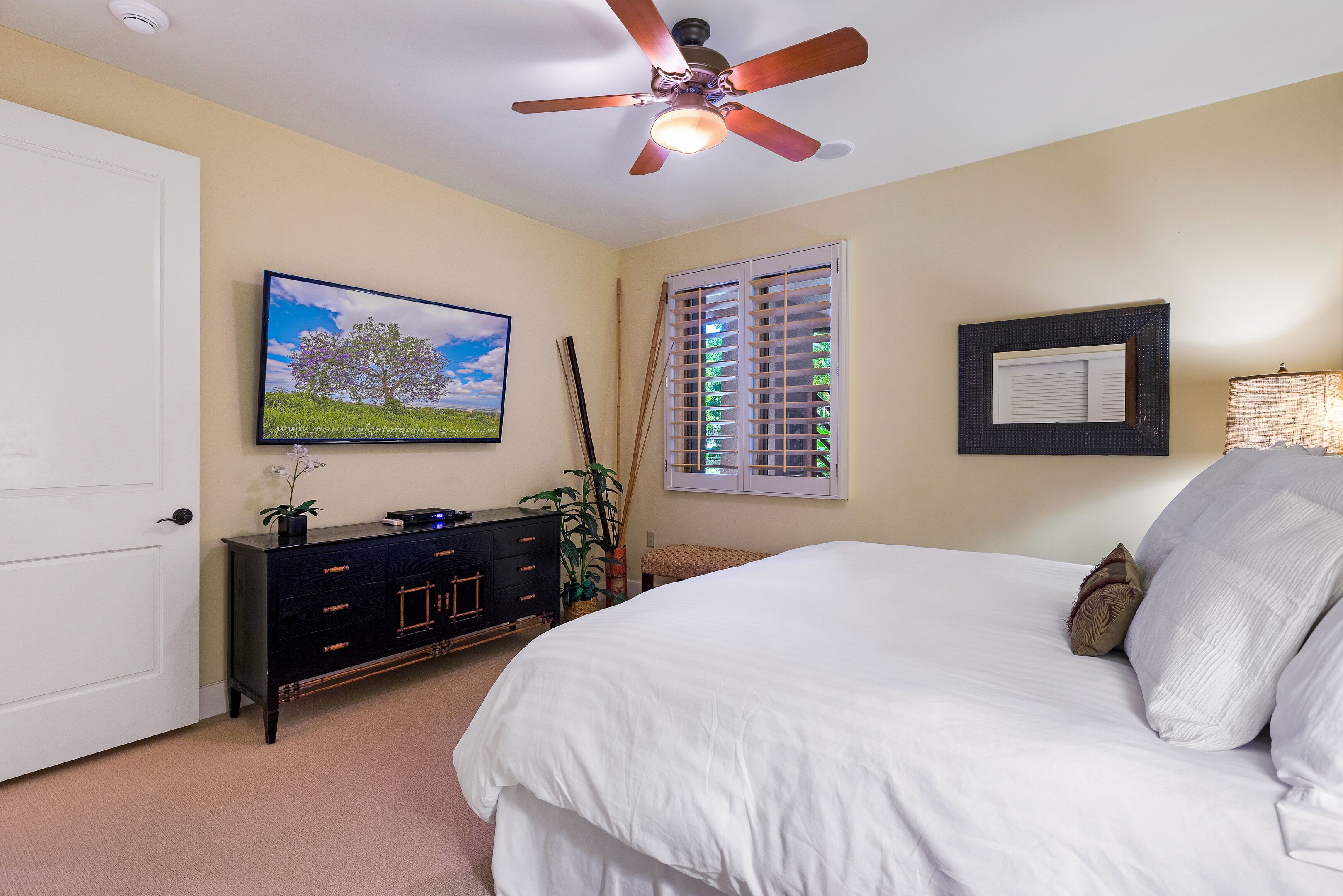 Quarto com cama, ventilador de teto, cômoda, televisão, janela com persianas e um quadro na parede.