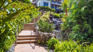 A lush garden with a waterfall, a stone staircase, and a building in the background. - The Maui Escape - Spectacular 2.5 Bdr Penthouse -Oceanfront Resort-Owner Direct! (Wailea)