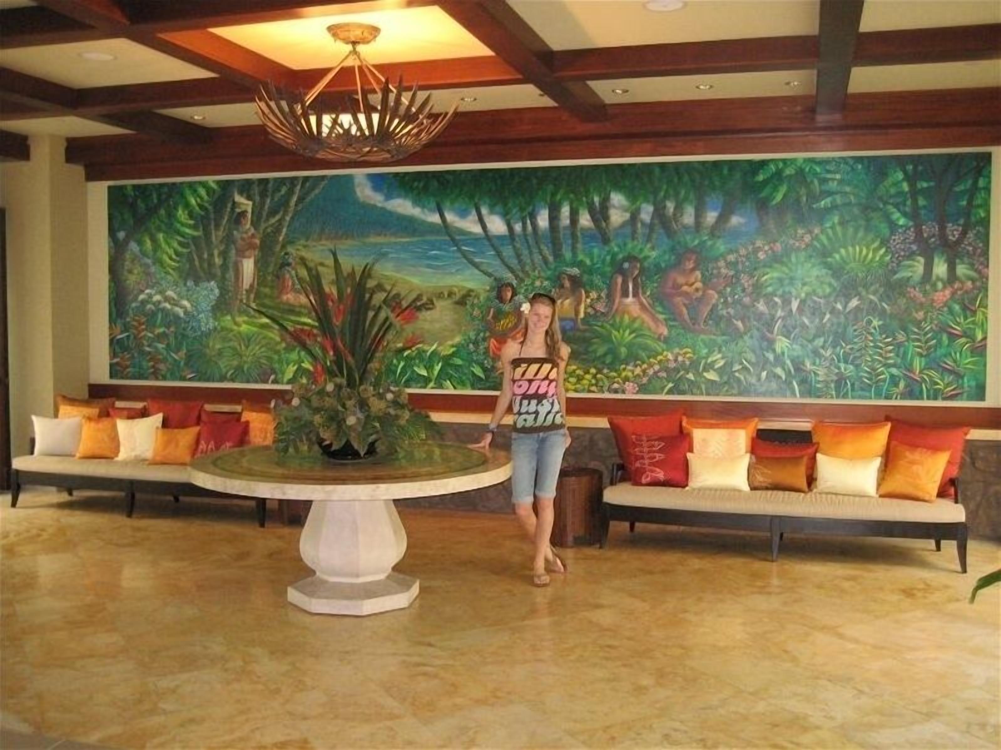 Eine Lobby mit einer großen Wandmalerei einer Dschungel Szene, einem runden Tisch mit einer Pflanze als Mittelpunkt und einer Sitzecke mit bunten Kissen.