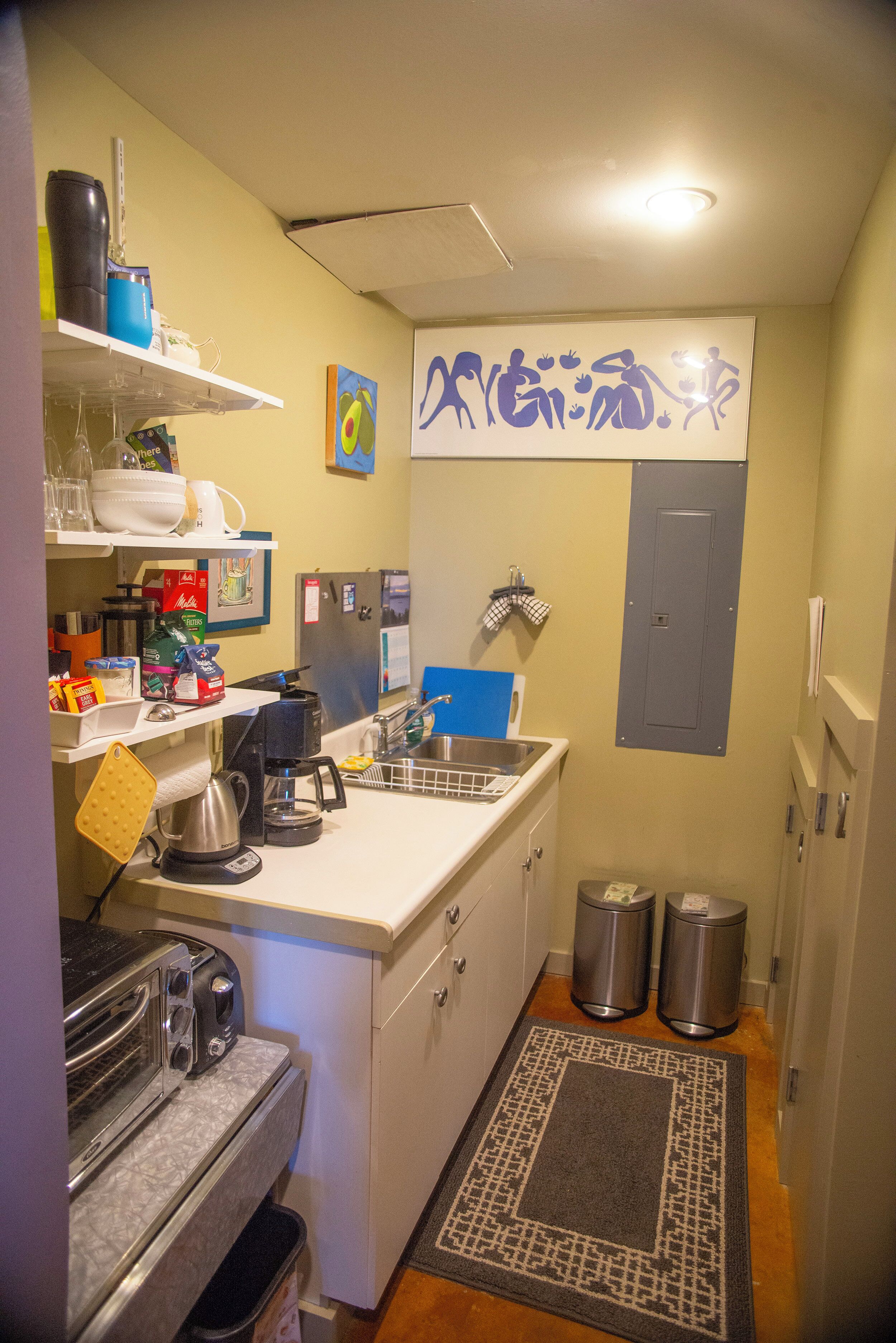 A compact kitchen with a coffee maker, toaster, and sink. There are two trash bins and a small rug on the floor.