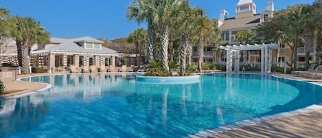 A large, clear swimming pool with a curved edge, surrounded by palm trees and a building with a dome roof.