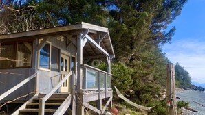 A wooden cabin with a staircase leading to a deck, surrounded by trees and a body of water.