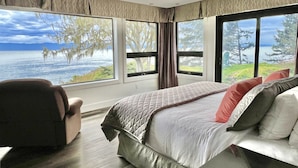 A bedroom with a large bed, a chair, and a view of a lake and trees.