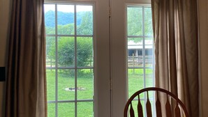 A room with a wooden table and a chair, a red patterned tablecloth, and a view of a green landscape through a door with curtains.