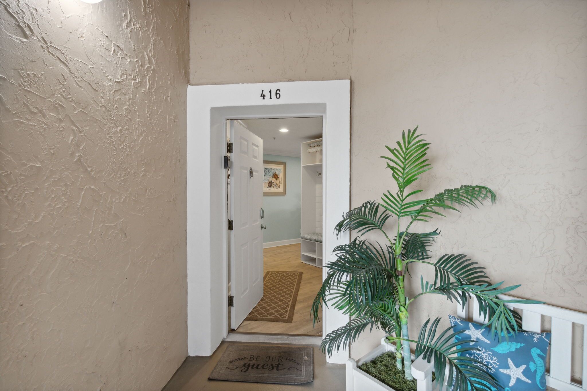 Un pasillo con una puerta identificada como '416' que da a una habitación con piso de madera y un tapete. En primer plano, hay una planta en maceta y un banco con cojín.